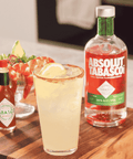 A glass of pale yellow cocktail on a wooden board, with a rim of reddish spice, beside a bottle of Absolut Tabasco rum and a small bottle of Tabasco hot sauce. A shrimp cocktail is blurred in the background.