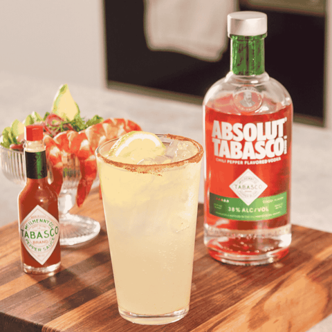 A glass of pale yellow cocktail on a wooden board, with a rim of reddish spice, beside a bottle of Absolut Tabasco rum and a small bottle of Tabasco hot sauce. A shrimp cocktail is blurred in the background.